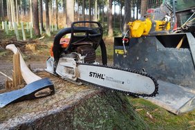 (c)pixabay.com/Mr_Neustadt Baumstumpf mit Hacke und Motorsäge. Dahinter Traktor.