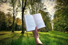 Hand hält offenes Buch, im Hintergrund Wiese und Bäume
